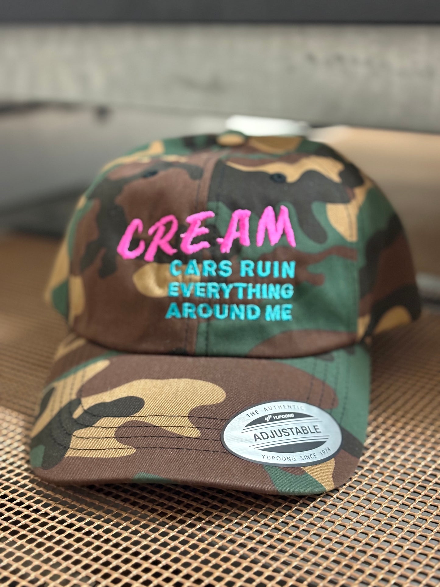 Camouflage cap with CREAM, cars ruin everything around me embroidered on it on a textured surface