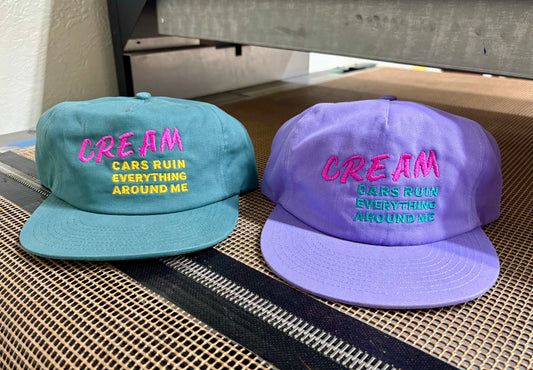 Two hats that say 'CREAM, Cars ruin everything around me', one blue and one purple, with text on a textured surface.